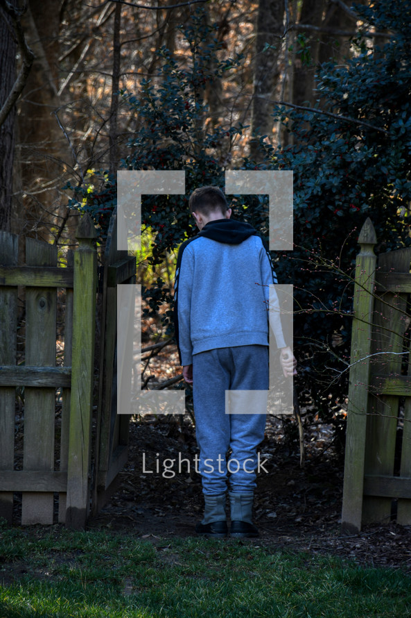 teen boy with a stick walking out a gate into a forest 