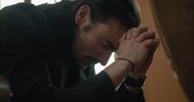 Young, emotional, anxious, and stressed man with long hair and black suit sitting in old church in worship and praying.