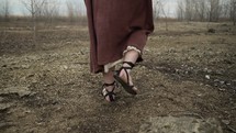 The feet of Jesus Christ, bible prophet (Noah, Moses, Elijah, John the Baptist, Abraham), or nomad traveler dressed in middle eastern, brown robes, sandals walking alone in cinematic, slow motion in the wilderness temptation by the Devil or Satan for 40 days and 40 nights.