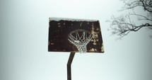 Old, rusty basketball hoop and court on snowy day. Slow motion snow and snowflakes fall in winter storm in small town.