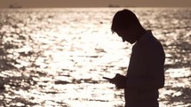 Guy looking at a cell phone in front of a body of water. 