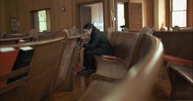 Young, emotional, anxious, and stressed man with long hair and black suit sitting in old church in worship and praying.