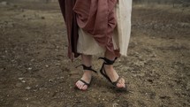 The feet of Jesus Christ dressed in sandals,  brown robes and tunic walking alone in cinematic, dramatic, slow motion in the wilderness temptation by the Devil or Satan for 40 days and 40 nights fasting and praying. Could also be used to depict a biblical prophet like Noah, Abraham, Elijah, Moses or John the baptist walking and traveling.