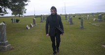 Sombre, sad young man in black suit walking in cinematic slow motion in cemetery through graveyard tombstones.