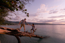 a couple holding hands standing on a fallen tree hanging over a shore 