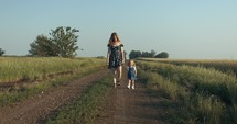 Mother and daughter walking on dirt road while holding hands in summer sunset.