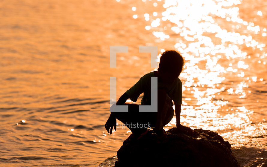 child standing on a rock at sunset along a shore 