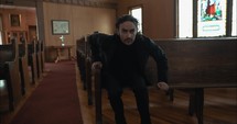 Young, emotional, anxious, and stressed man with long hair and black suit sitting in old church in worship and praying.