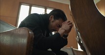 Young, emotional, anxious, and stressed man with long hair and black suit sitting in old church in worship and praying.