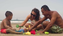 happy couple making sand castle at tropical beach with their son. Child with mom and dad outdoor at seaside playing together