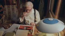 Elderly white-haired writer with cigarette in his mouth typing on red typewriter and revising handwritten notes scattered on desk illuminated by lamp in cozy room filled with books. High angle view
