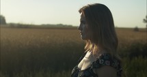 Sunlight shines on young woman standing outside watching sunset in wheat field.