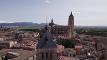 Establishing aerial view across the rooftops of San Estaban church and Segovia cathedral with mountain landscape skyline	