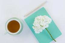 flowers on a turquoise journal, pencil, and cup of tea on a table 