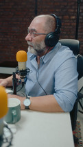 Senior journalist in headphones sitting at desk with microphone and recording podcast with guest at studio. Vertical format footage