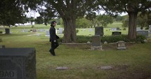 Sombre, sad young man in black suit walking in cinematic slow motion in cemetery through graveyard tombstones.