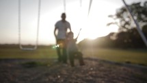 Silhouette of a happy father and son. Dad pushes his young boy on a swing at a playground in the sunrise or sunset sunlight in cinematic slow motion. 