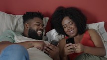Happy young couple in bed on their phone on social media, browsing internet and online meme. Black couple in bedroom with smartphone