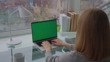 Woman typing on laptop with green screen sitting at the desk home office with big window slow motion
