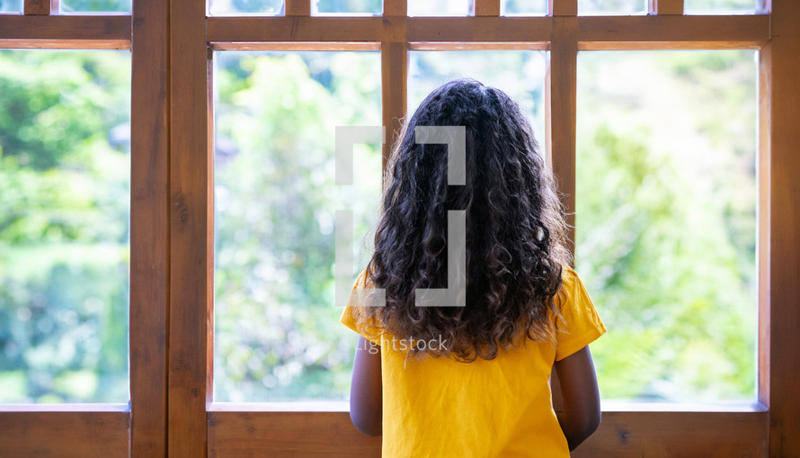the back of a young girl looking out a window on a bright, sunny day 