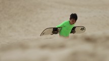Boy walking sandboard up desert dunes on summer day - wide shot