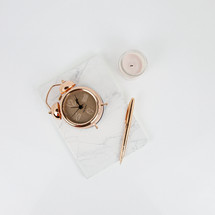 rose gold, alarm clock, flower, notebook, pen, and candle on white desk 