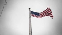 Tattered and worn American, USA flag blowing in cinematic slow motion in the wind in front of grey cloudy sky.