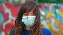 a young woman wearing a face mask standing in front of a graffiti covered wall 