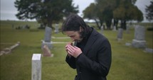Young, sad man in black suit crying in cemetery at graveyard tombstone grieving in cinematic slow motion.