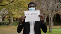 a man in a face mask holding a 6 feet apart sign 