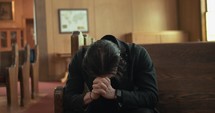 Young, emotional, anxious, and stressed man with long hair and black suit sitting in old church in worship and praying.