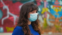 a young woman wearing a face mask standing in front of a graffiti covered wall 