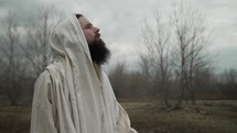 Religious Christian man, bible prophet or Jesus Christ with beard wearing a white tunic lifts hands to sky praying in worship to God.
