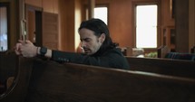 Young, emotional, anxious, and stressed man with long hair and black suit sitting in old church in worship and praying.