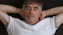 Gray haired man lying down with his eyes closed.