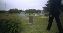 Young, sad man in black suit kneeling in cemetery at graveyard tombstone grieving in cinematic slow motion.