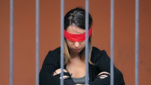 Woman with a red blindfold covering her eyes sitting behind metal bars.