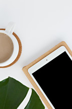 tropical plant leaf, coffee cup, iPad, and notebook on a white desk 