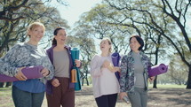 Group of positive female friends in sportswear holding exercise mats and water bottles, smiling and chatting while walking together in the park after fitness class