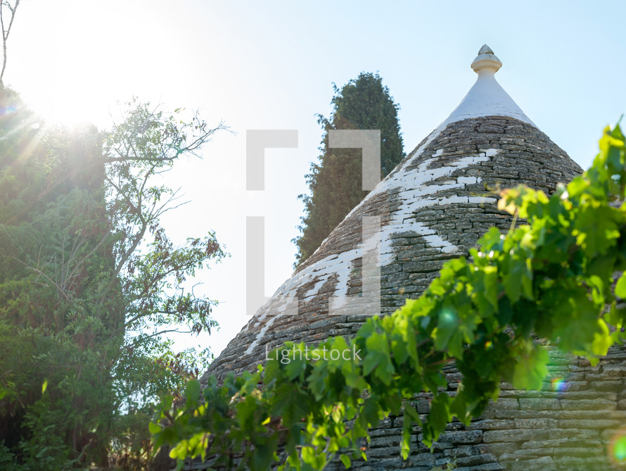 Sunlight shining through trees over a trullo in alberobello, a traditional apulian dry stone hut with a conical roof