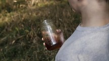 A young man in the woods holding a jar of alcohol. A teen boy standing in the woods in slow motion.