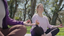 Mid-aged woman sitting cross legged on exercise mat, looking at yoga teacher, then closing her eyes and holding mudras during meditation in the park