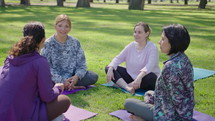 Group of mature female friends in sportswear sitting on exercise mats on green lawn in the park, smiling and talking while resting after outdoor yoga practice