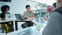 Young gen Z man answering questions of colleagues and explaining financial report, siting with business team around meeting table in modern office
