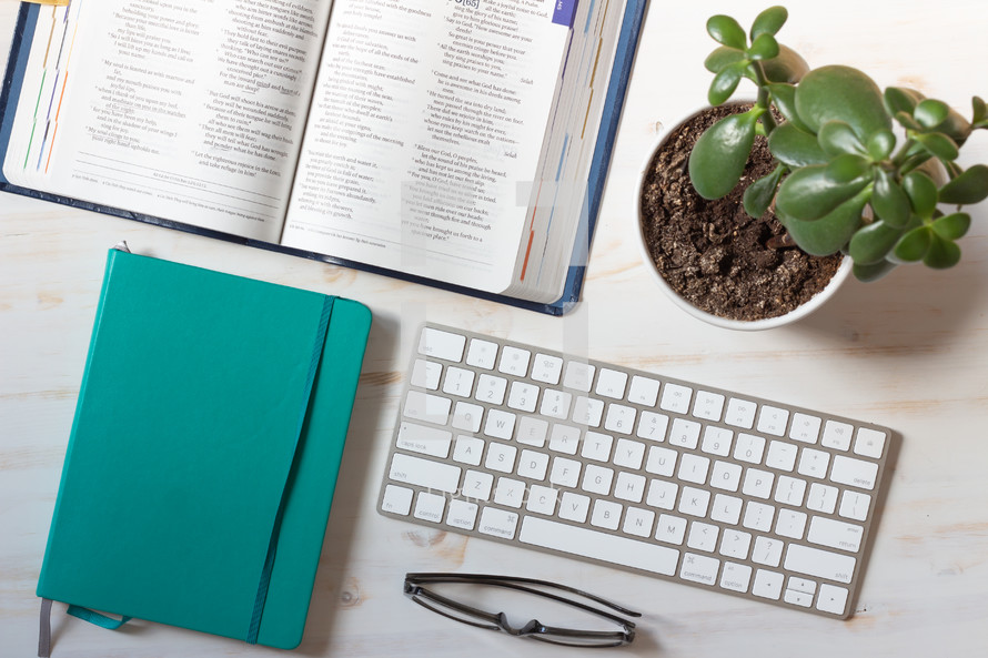 Bible, journal, computer keyboard, reading glasses, and houseplant on a desk 