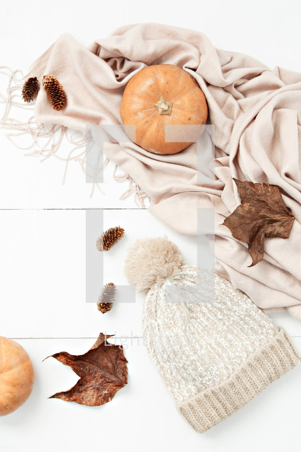 scarf, pumpkin, fall leaves, pine cones, and beanie on a white wood background 