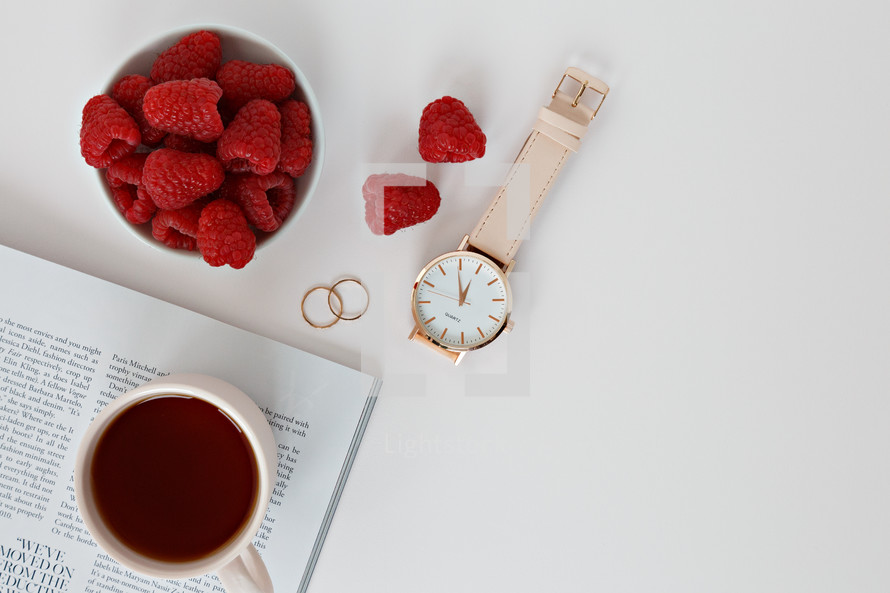 Raspberries in a bowl, watch, magazine, rings, and coffee cup 