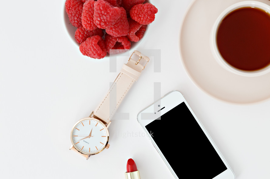 Raspberries in a bowl, iPhone, lipstick, watch, magazine, rings, spoon, and coffee cup