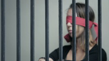 Woman with a red blindfold covering her eyes sitting behind metal bars.