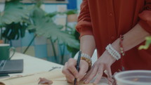 Woman attaching dried leaf to page with tape, then writing its name while making herbarium book at desk in indoor home garden. Close-up view, tilt-up shot
